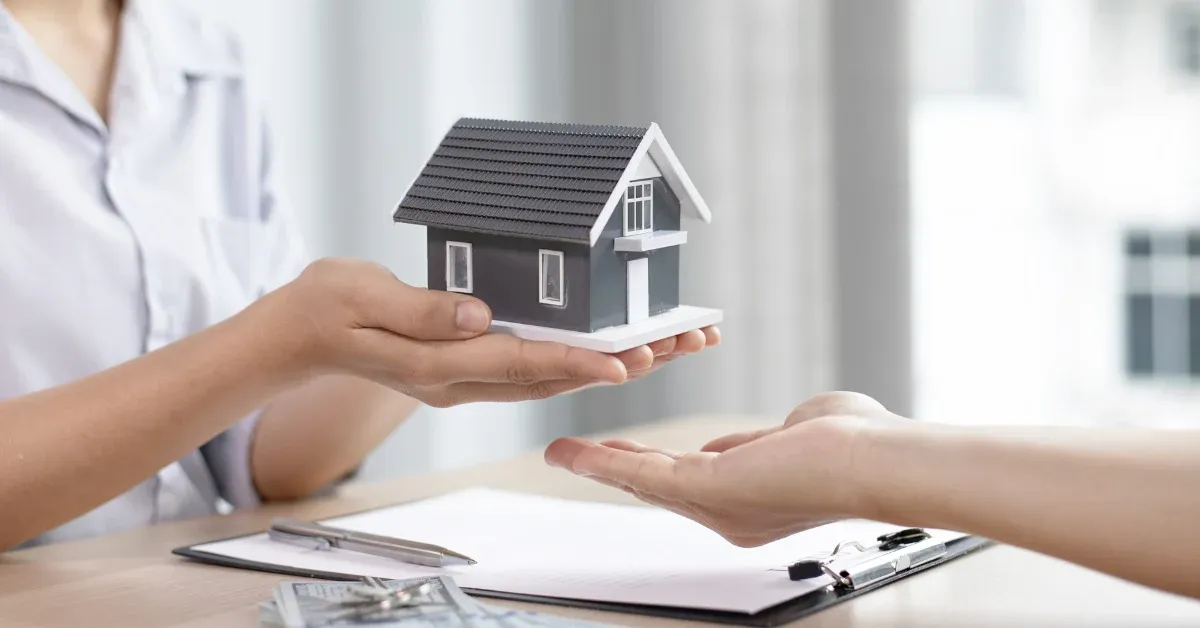 One person handing a small model house to another over a signed document